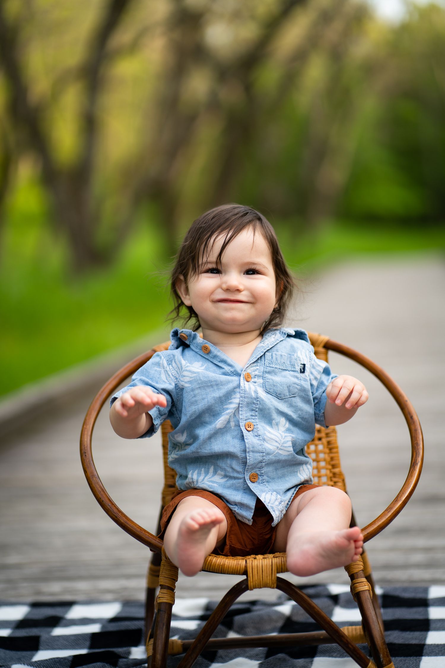 Small child sitting in wicker chair on checkered blanket along wooded path.