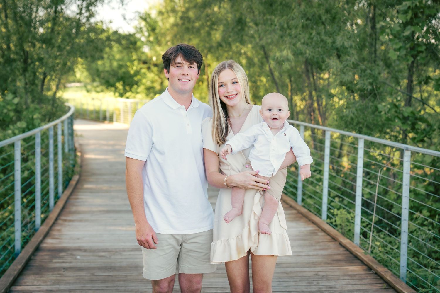 Family in white and neutral clothing poses on green metal bridge in natural setting.