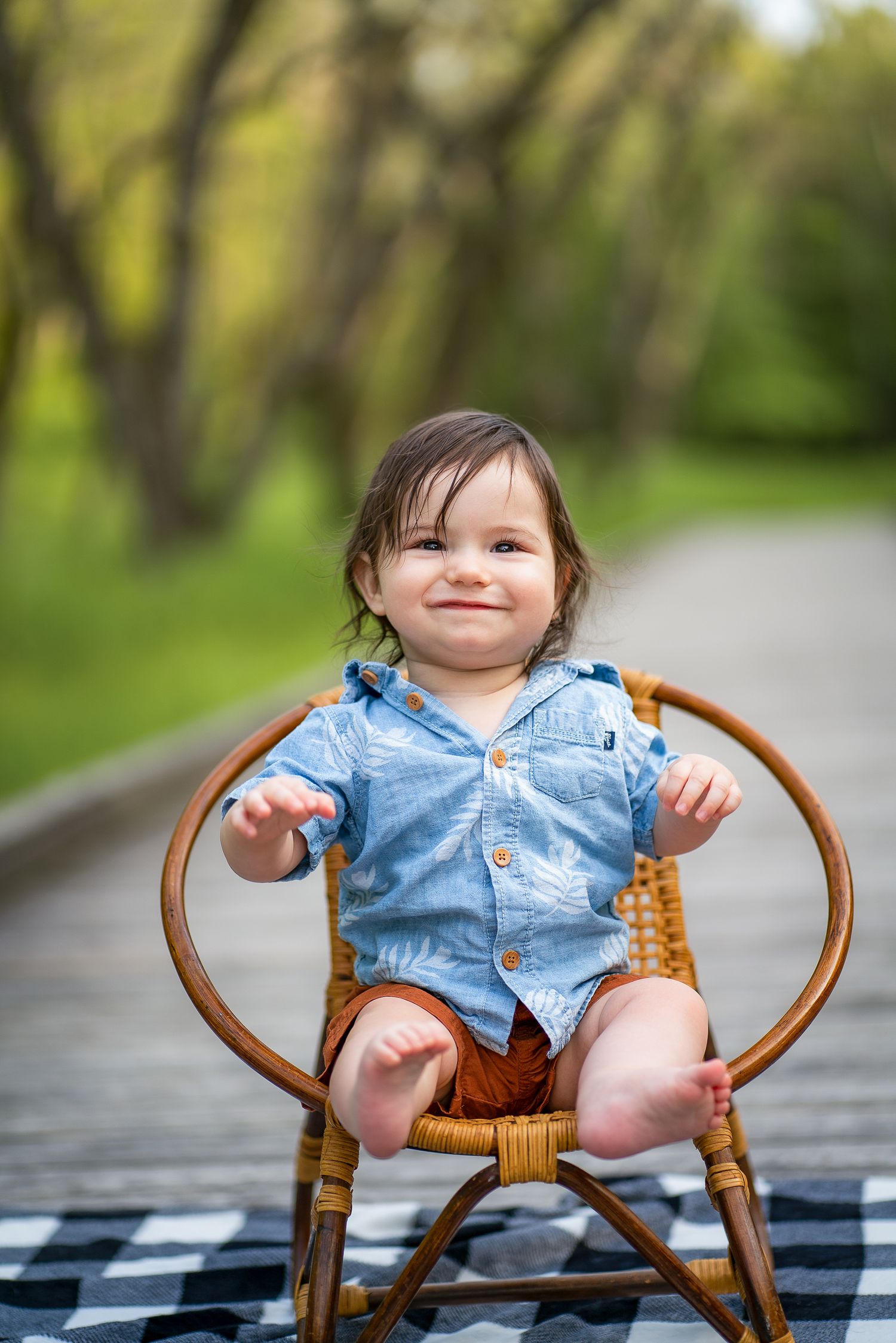 A small child wearing denim sits in a wooden chair outdoors on a sunny day with trees in the background.