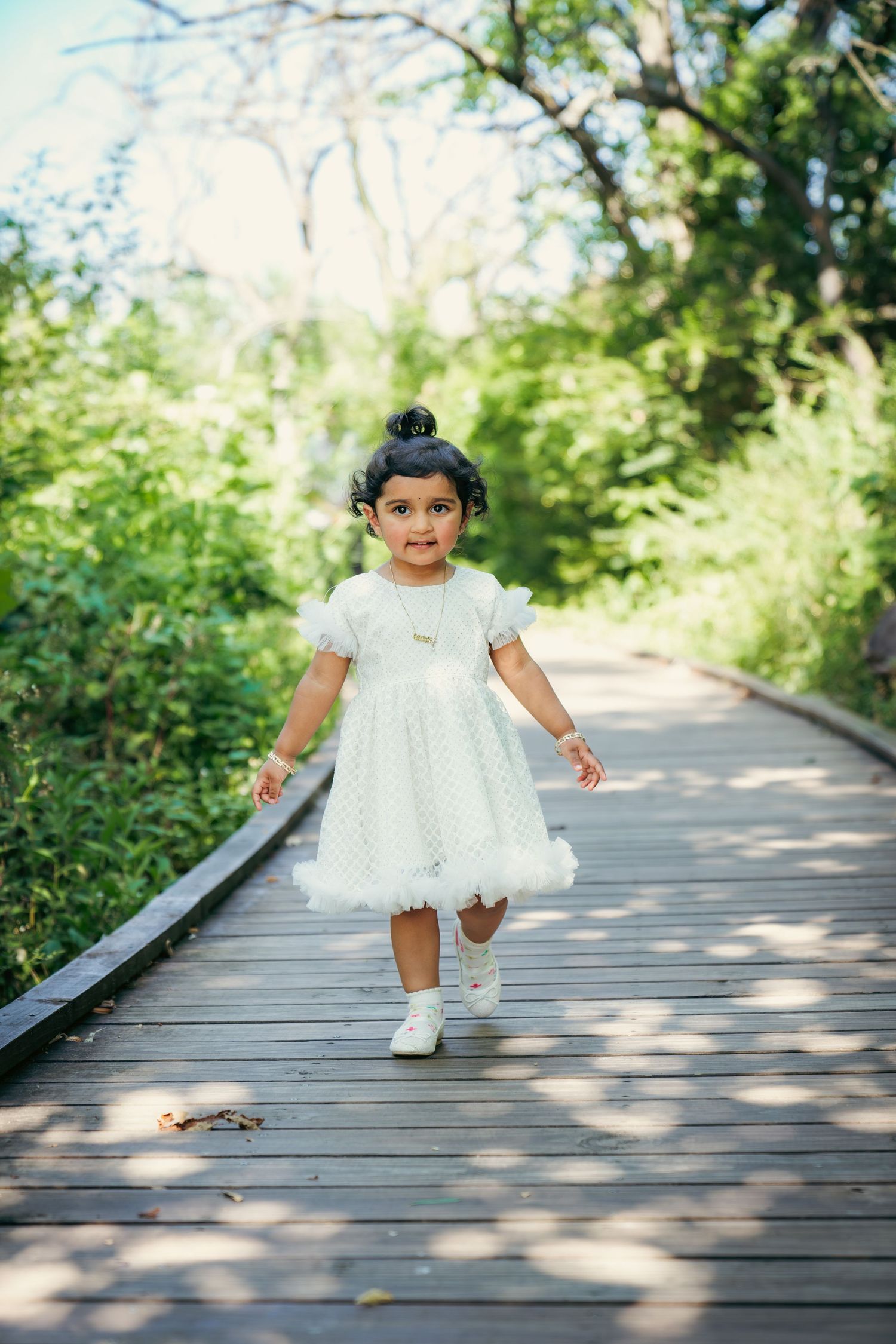 A young child wearing a white dress crouches down playfully on a sunlit wooden pathway.