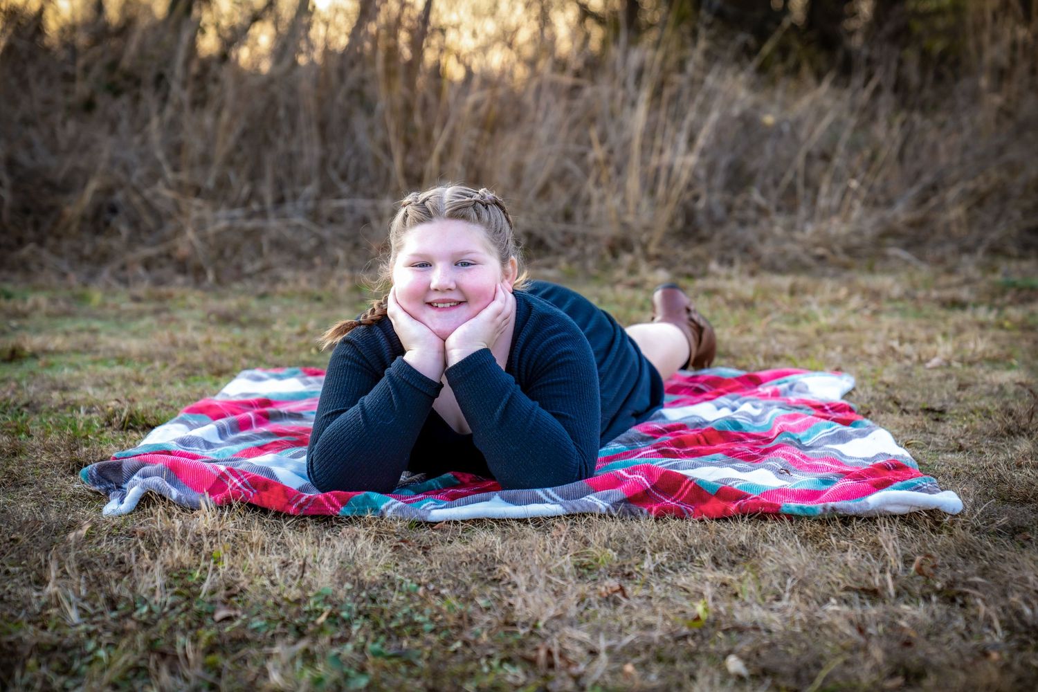 A person lies on a red and white checkered blanket in a grassy field at sunset.