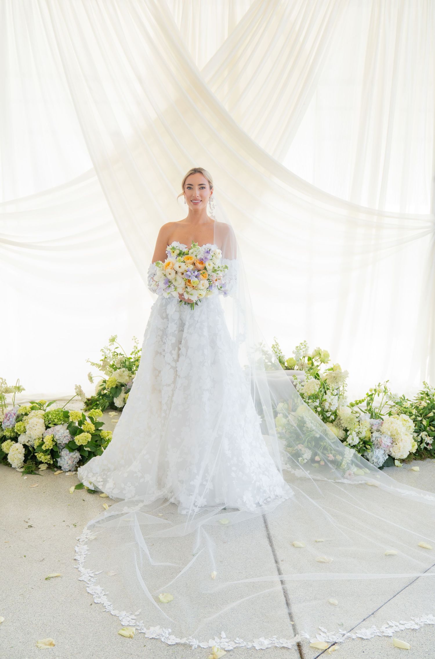 Full-length bridal portrait with cathedral veil and pastel bouquet in white draped setting.
