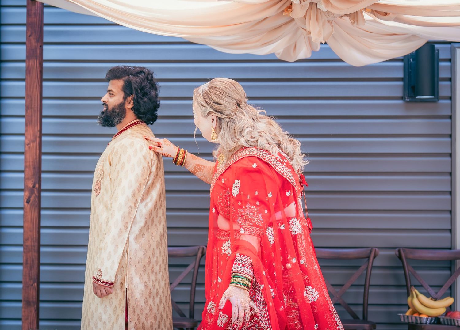 A couple in traditional Indian wedding attire pose under a draped canopy with peach fabric and floral decorations.