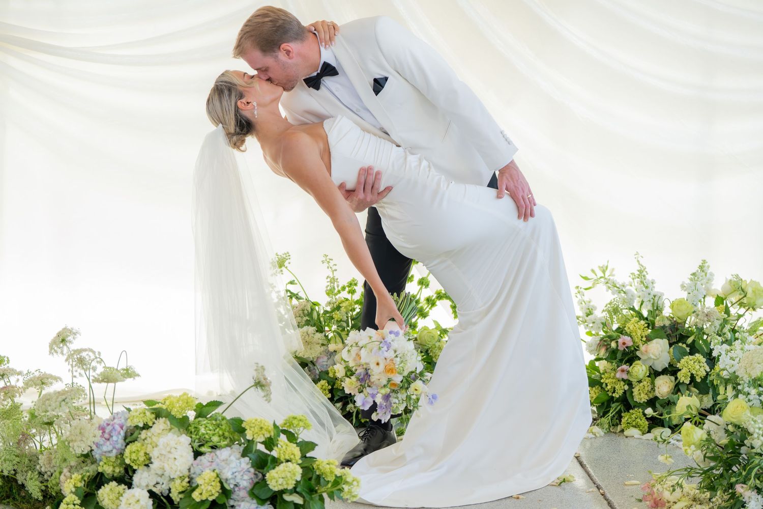 Wedding couple sharing a romantic dip kiss pose surrounded by white floral arrangements.