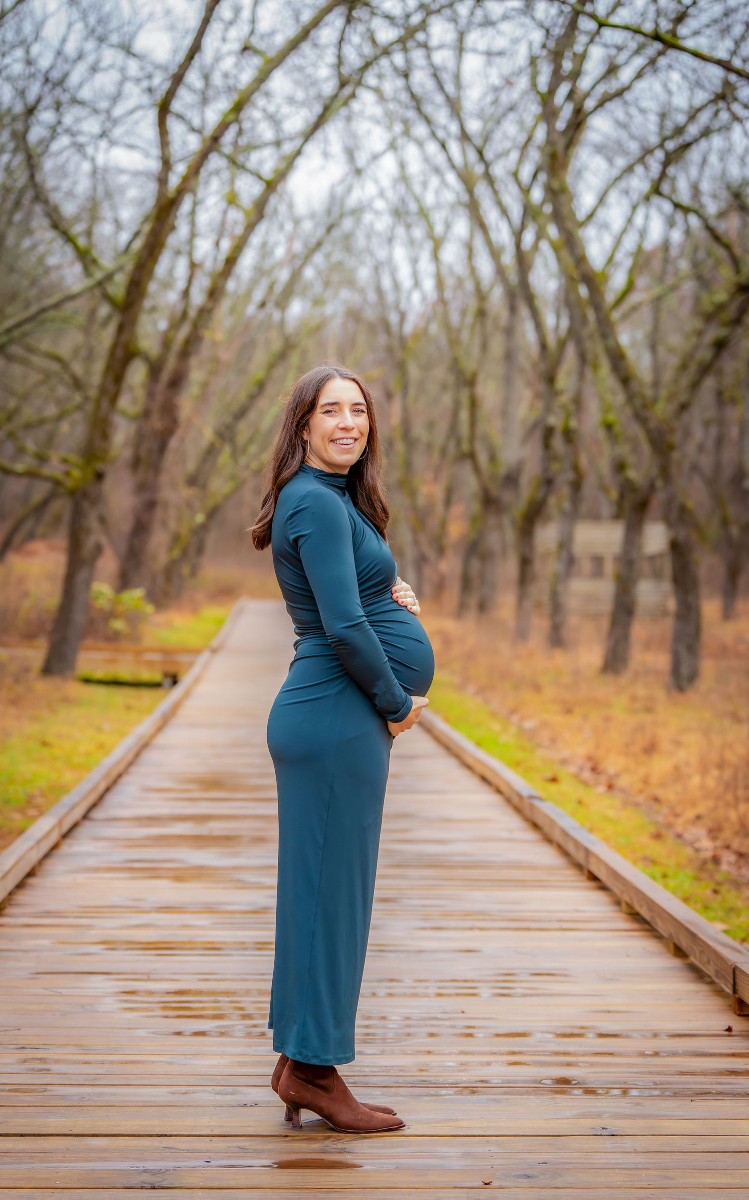 A woman in a teal dress poses on a wooden boardwalk surrounded by autumn trees.