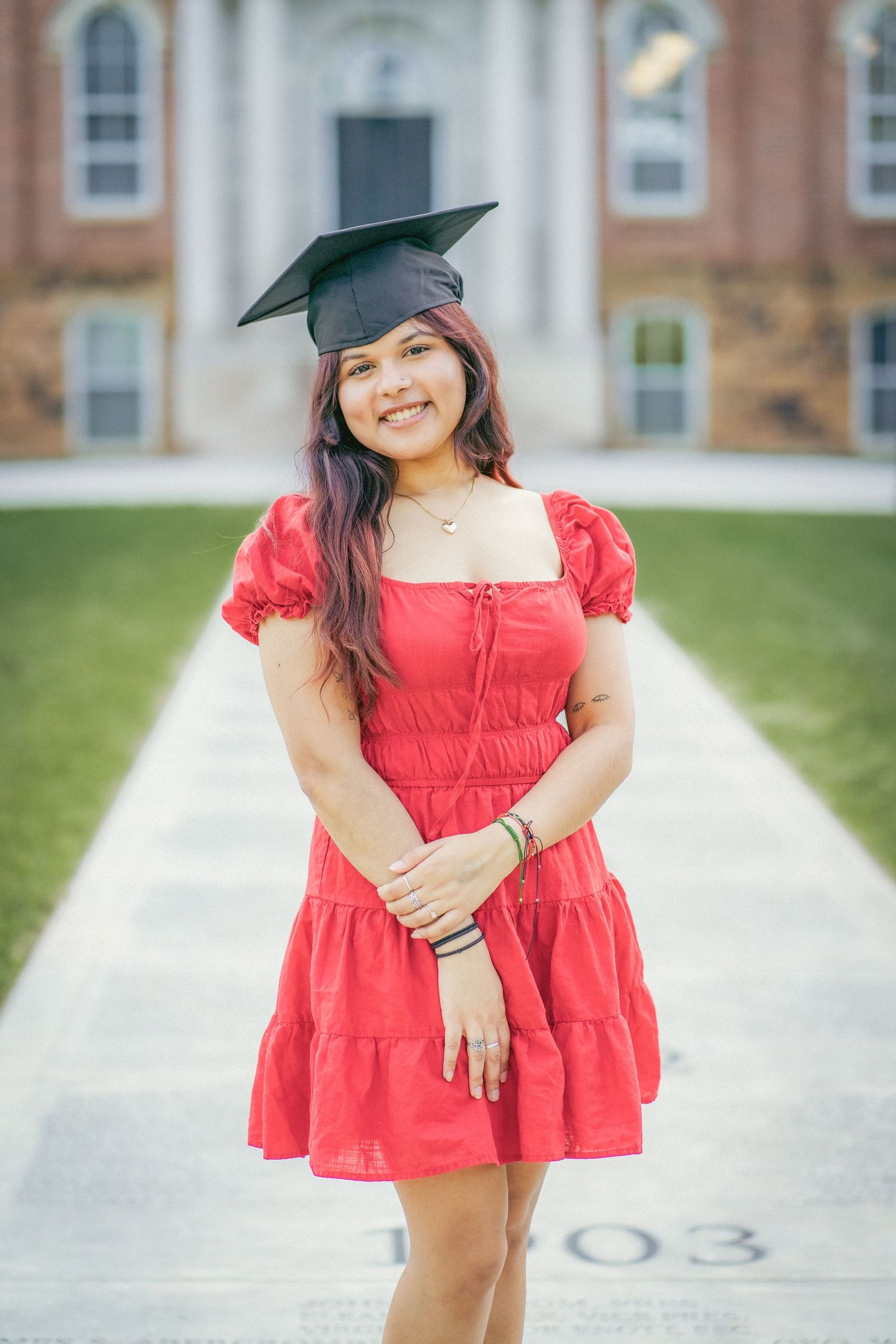 Graduate in red dress and cap posing in front of classic university architecture.