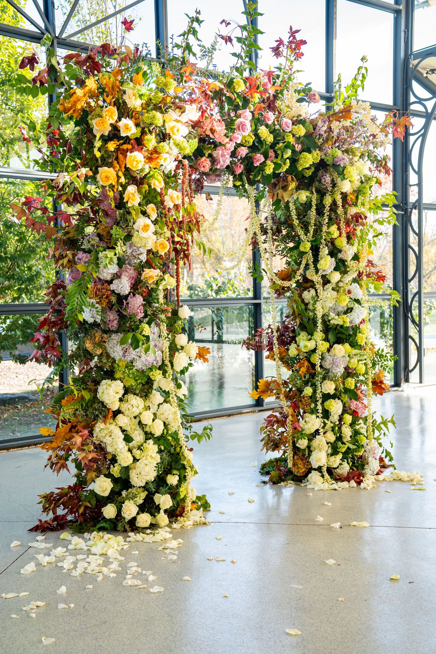 Luxurious wedding arch decorated with colorful autumn flowers and greenery in a glass-walled venue.