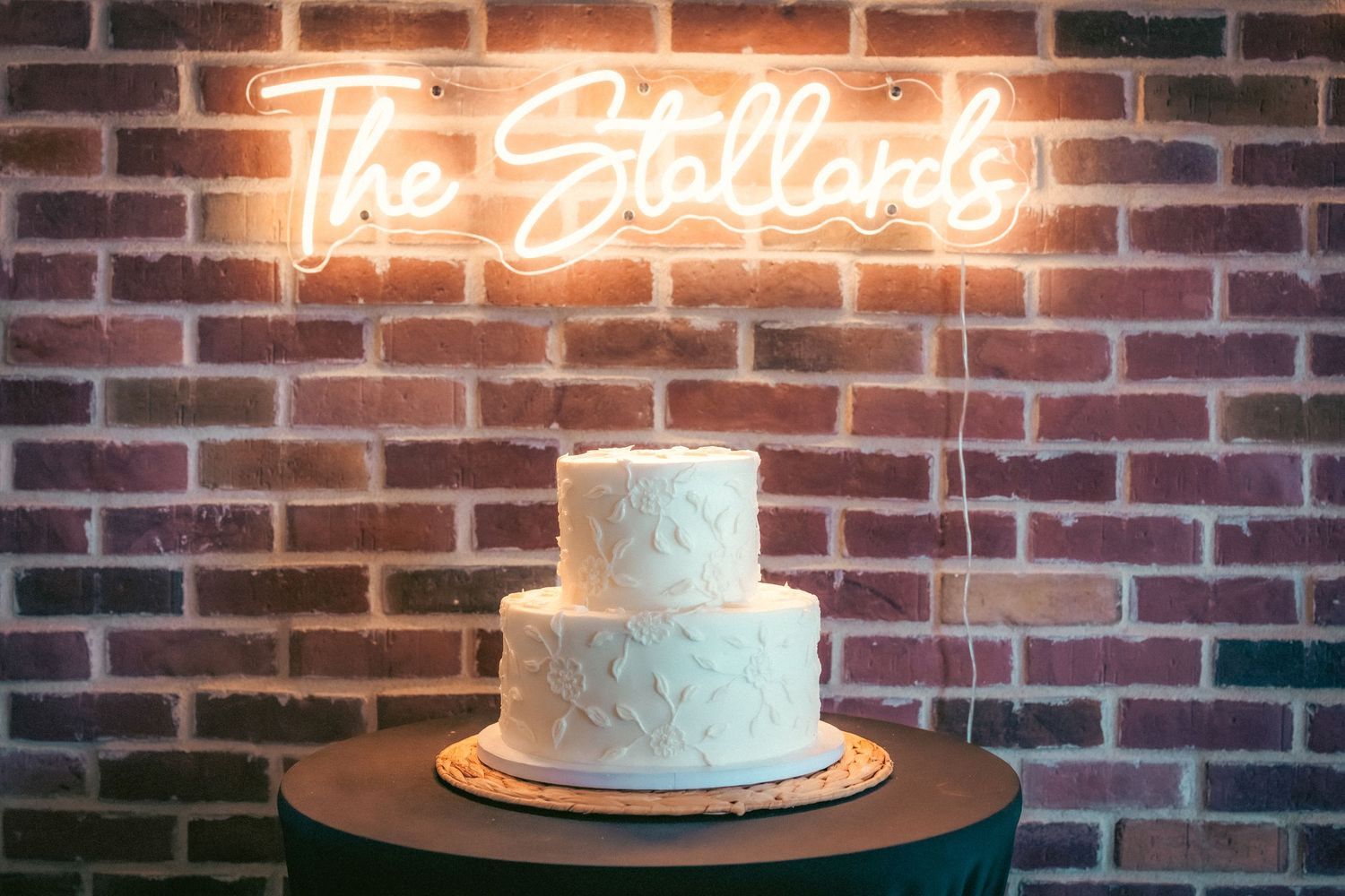 White wedding cake displayed in front of illuminated neon signage on exposed brick wall.