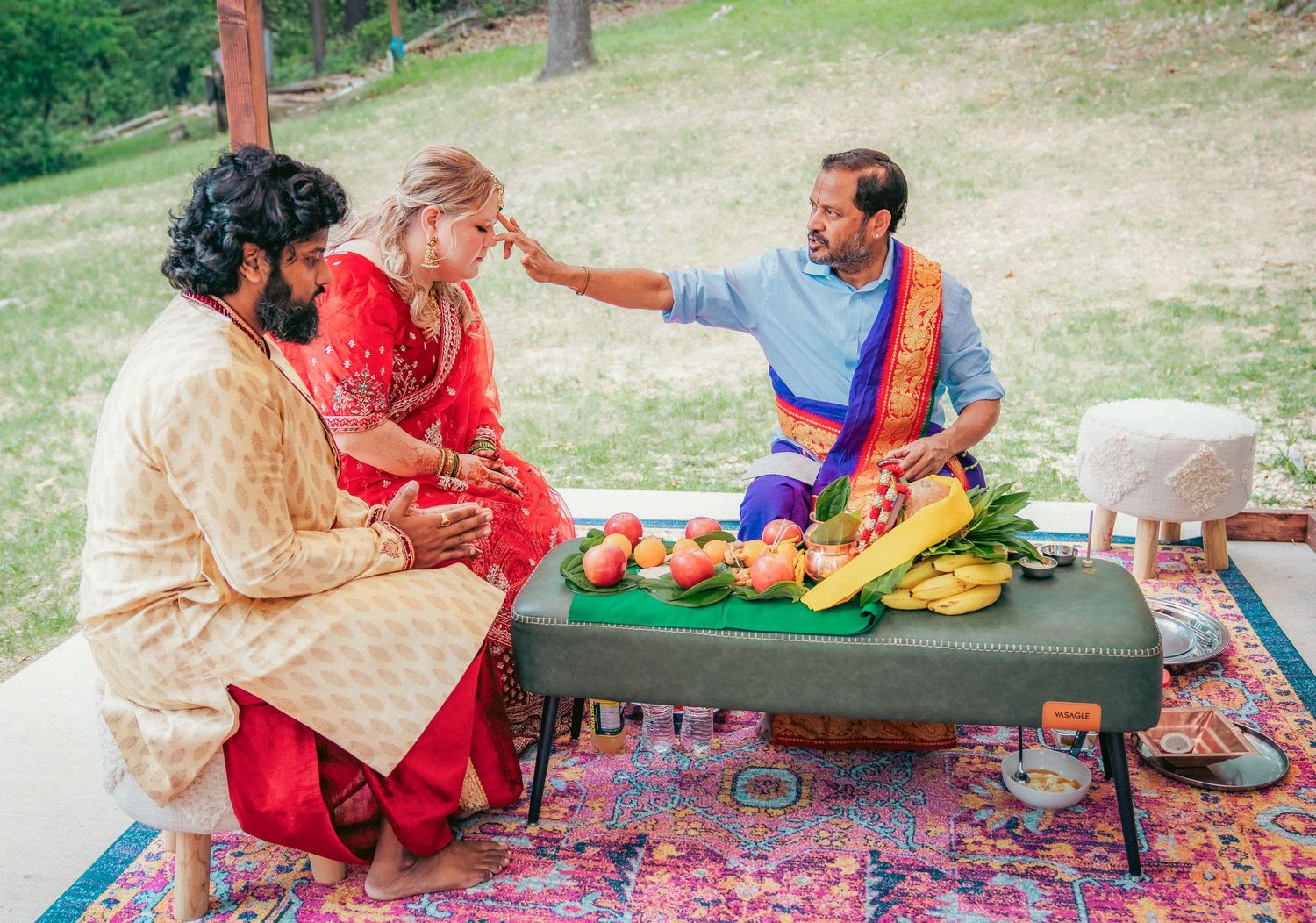 Wedding guests participate in traditional Indian ceremony rituals on colorful rugs under a canopy.