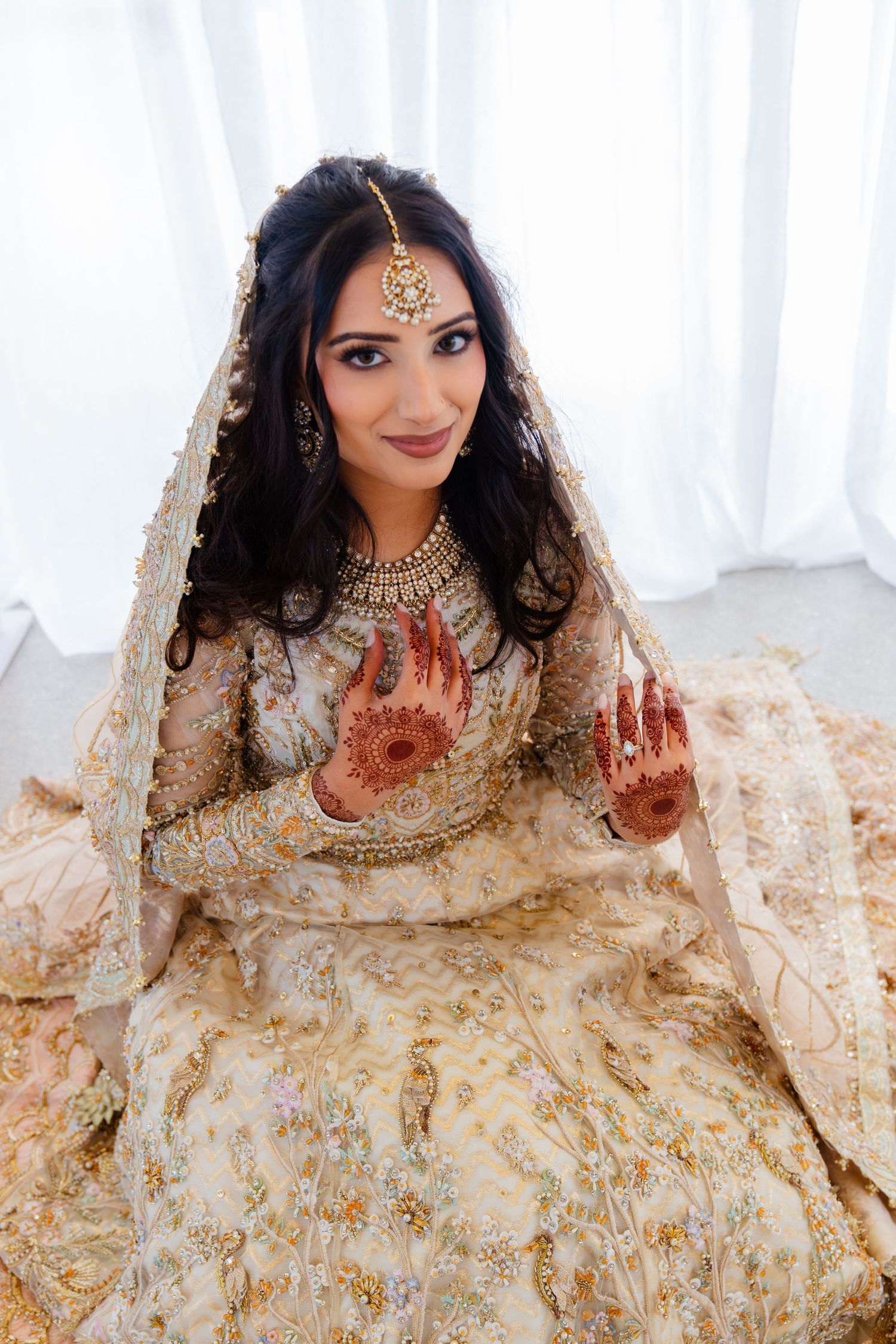 Traditional South Asian bride in cream and gold lehenga showcasing intricate henna designs on her hands.