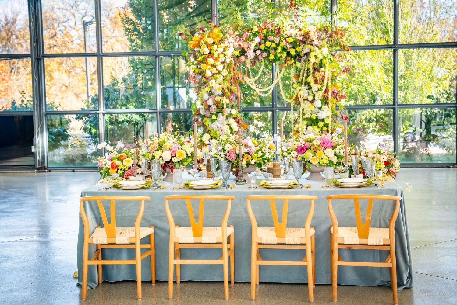 Elegant wedding reception tables set up in a bright sunlit venue with autumn foliage visible through windows.