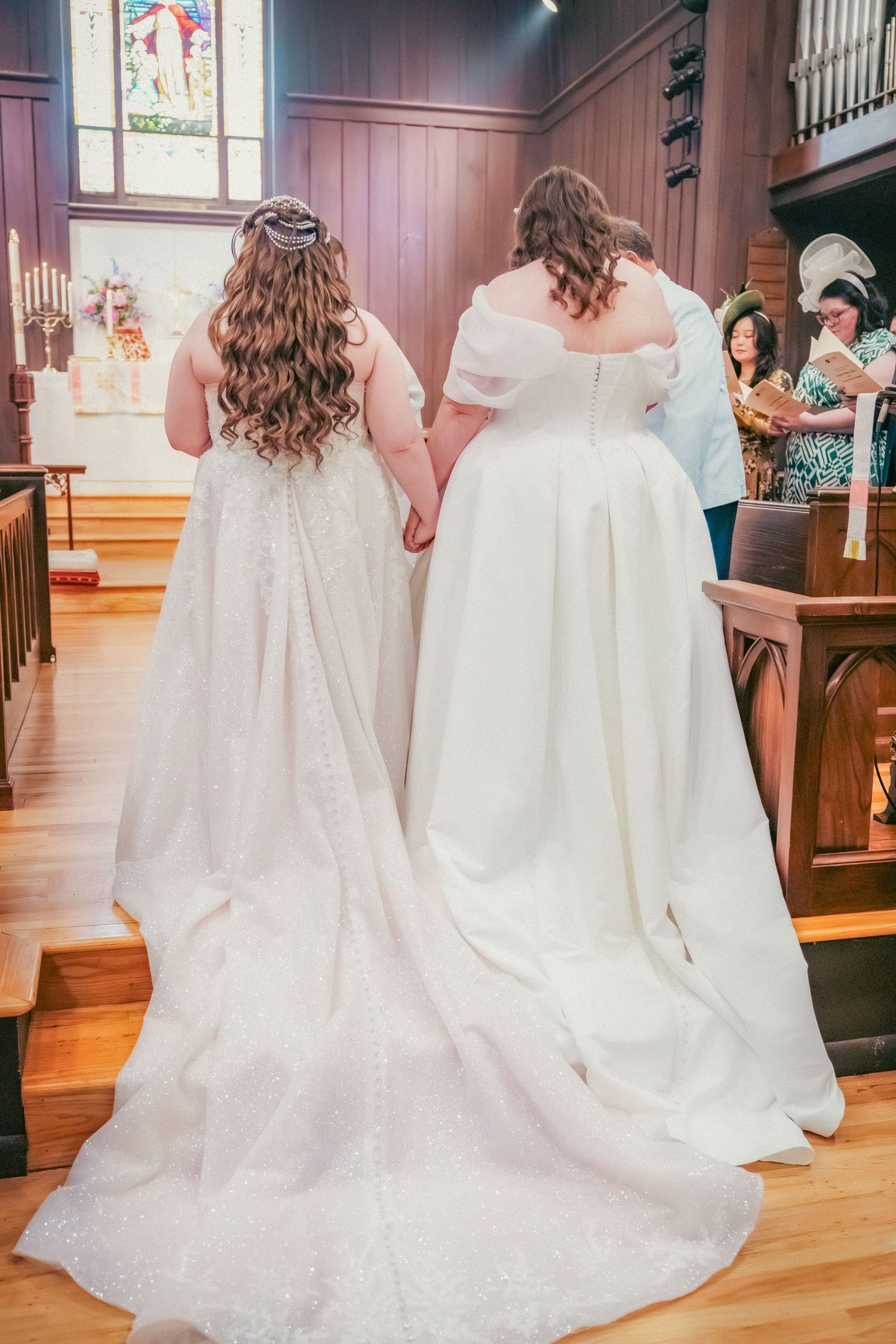 Back view of wedding dresses with long trains in a church setting with stained glass windows.
