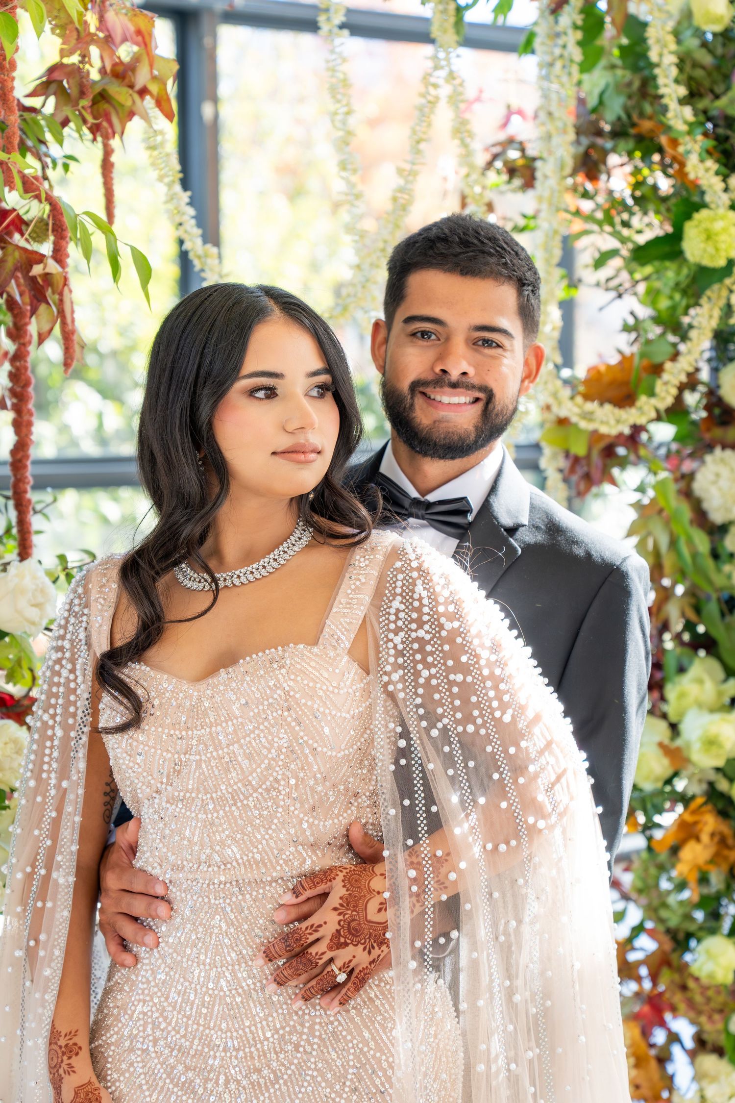 A couple poses in elegant wedding attire under a floral arch, with the bride wearing a sparkly beaded gown.