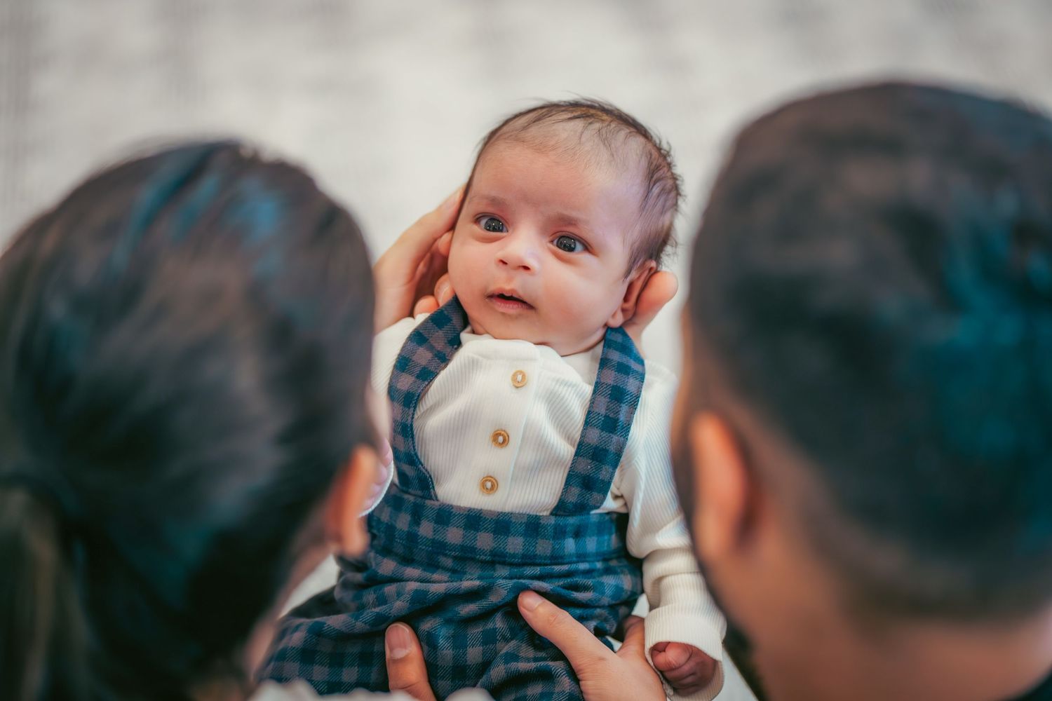 Baby wearing blue plaid overalls looking at camera.