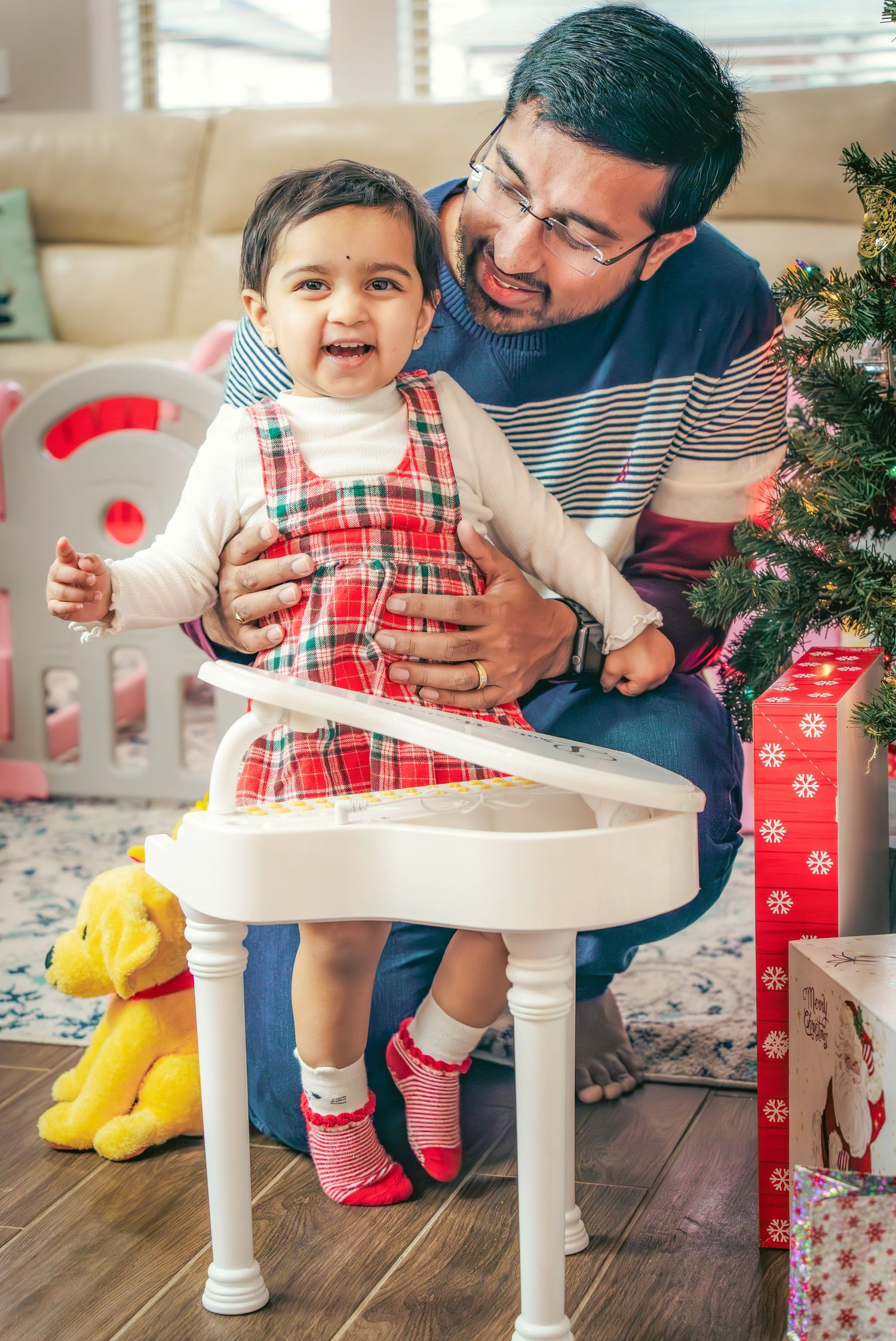 Holiday scene with a small child in a plaid dress and yellow boots standing at a white table near a Christmas tree.