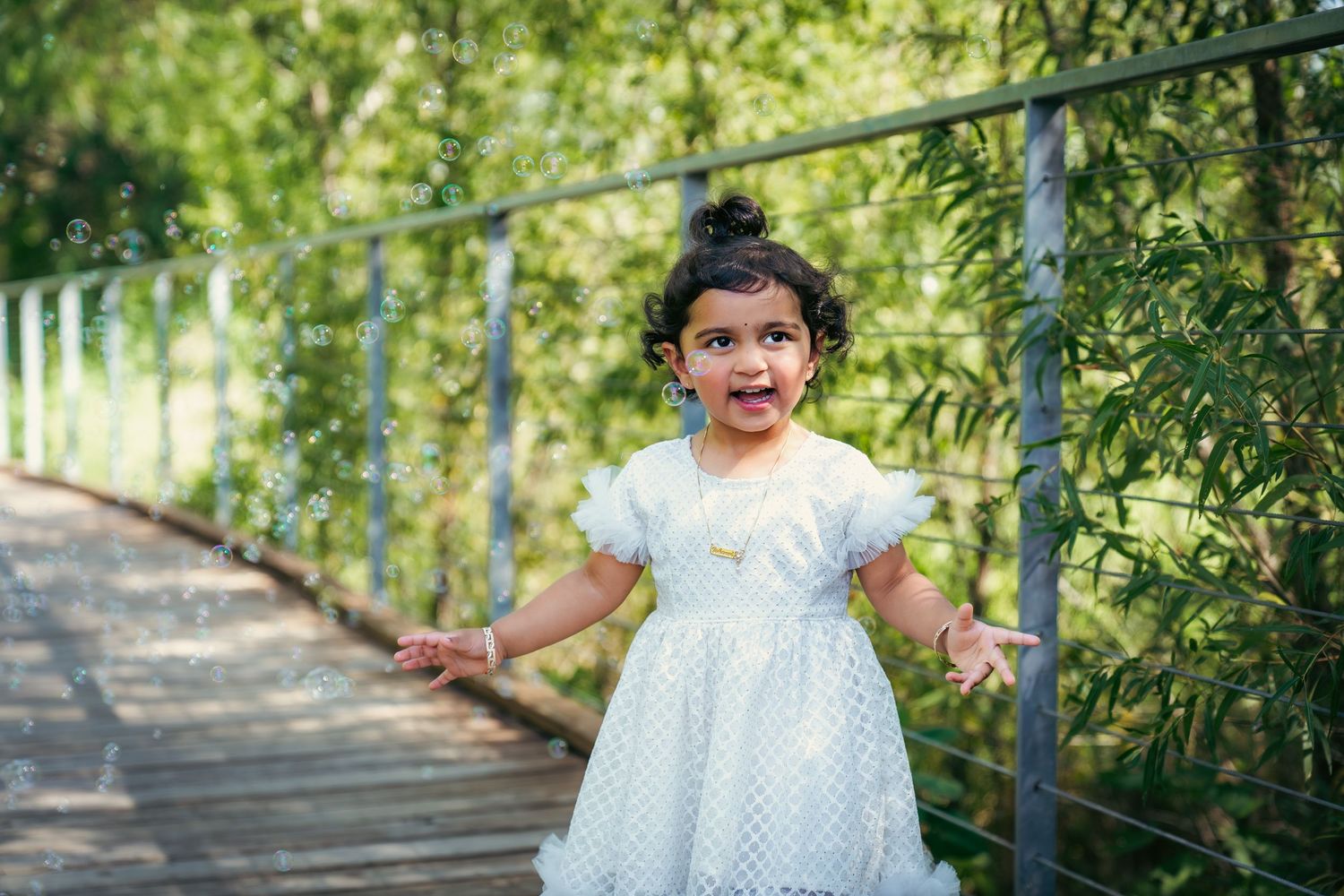 A child in a white dress plays energetically on a wooden bridge surrounded by trees.