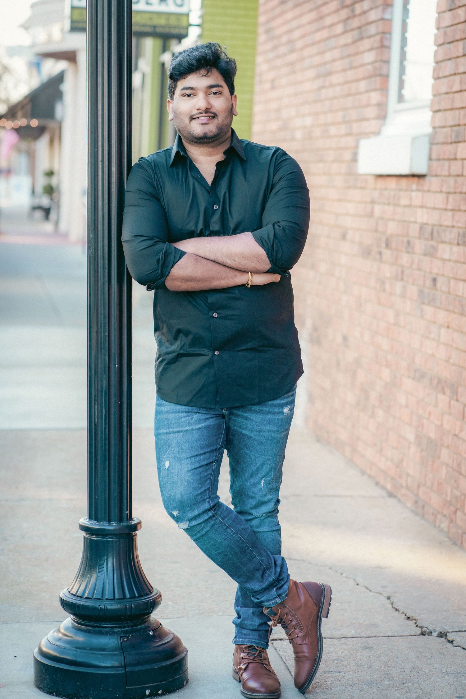 A person in a black shirt and jeans poses casually against a brick wall with decorative lamp post.