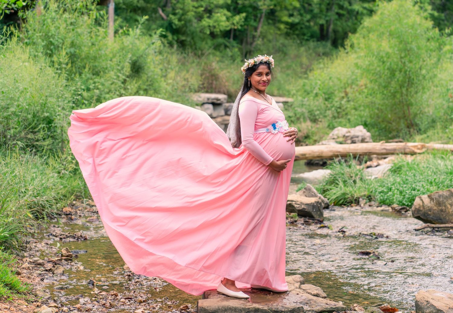 A pink dress flows dramatically against a natural creek setting with green foliage.
