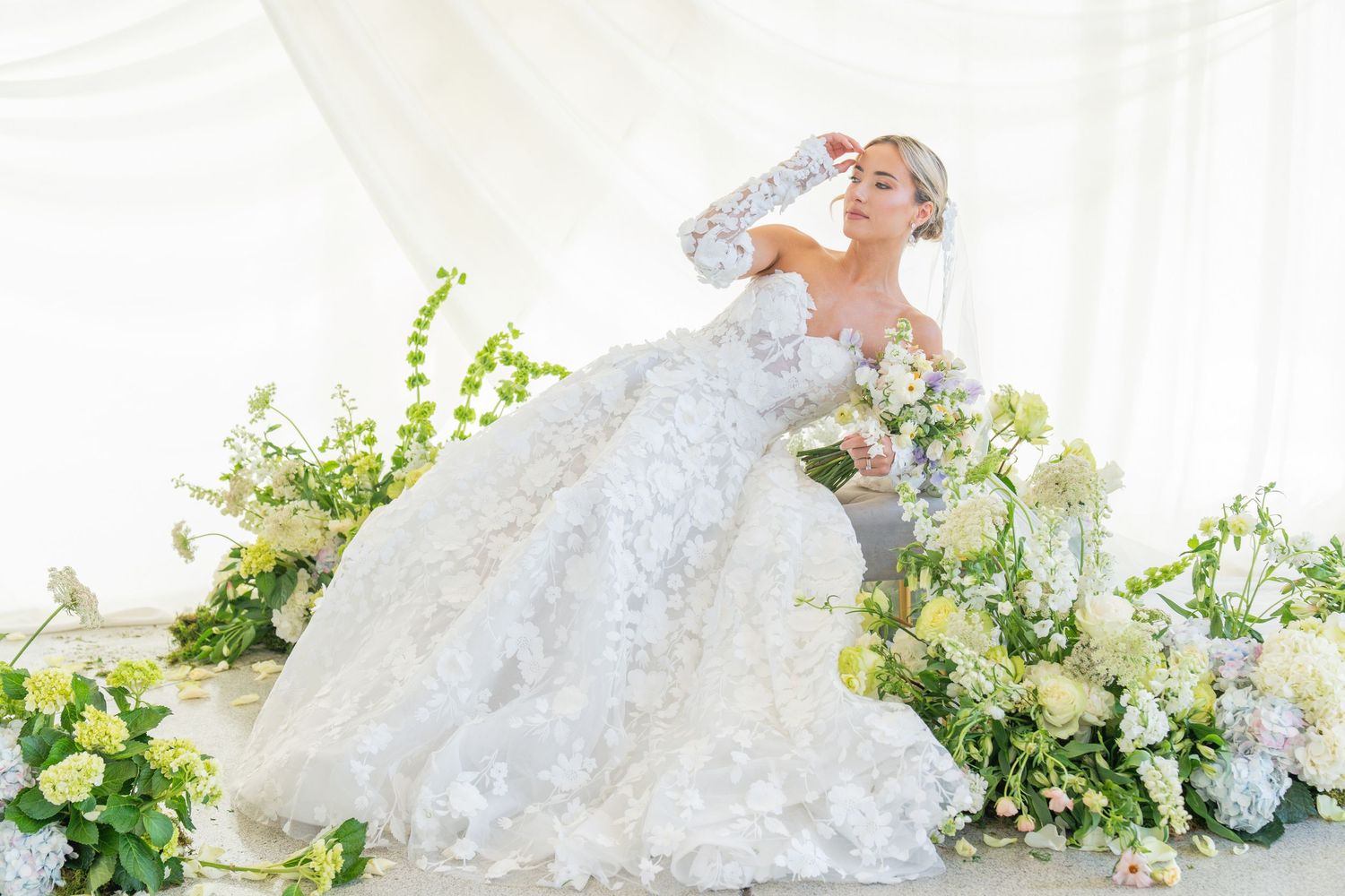 Romantic bridal portrait with white lace gown amid green and white floral arrangements.