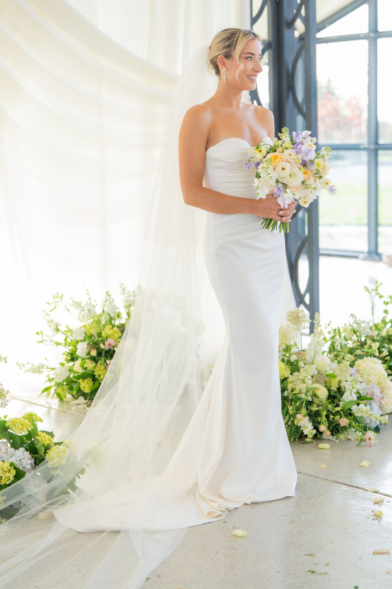Bride in white gown poses gracefully with floral bouquet in sunlit tent setting.