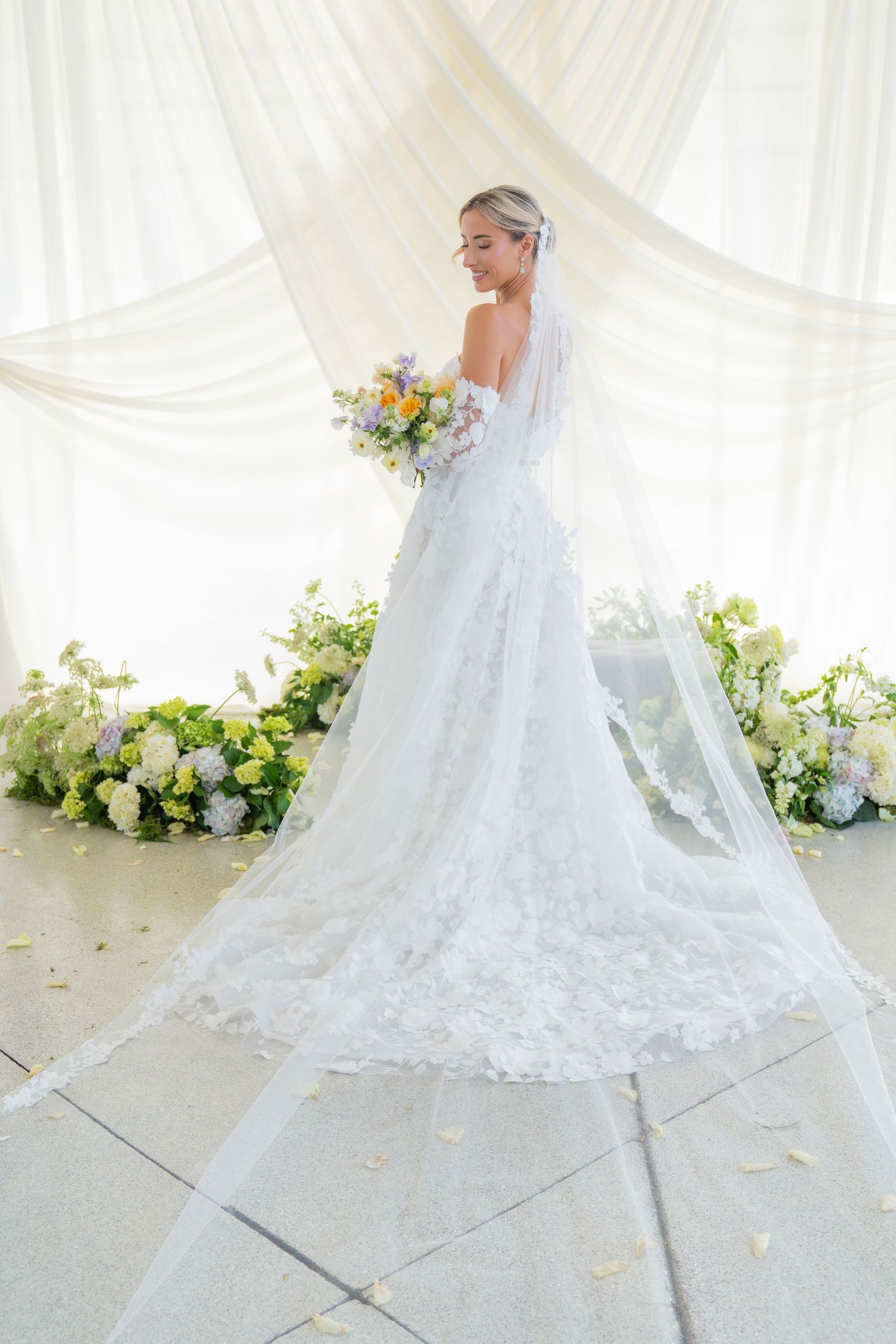 Back view of lace wedding gown with cathedral train surrounded by floral arrangements.