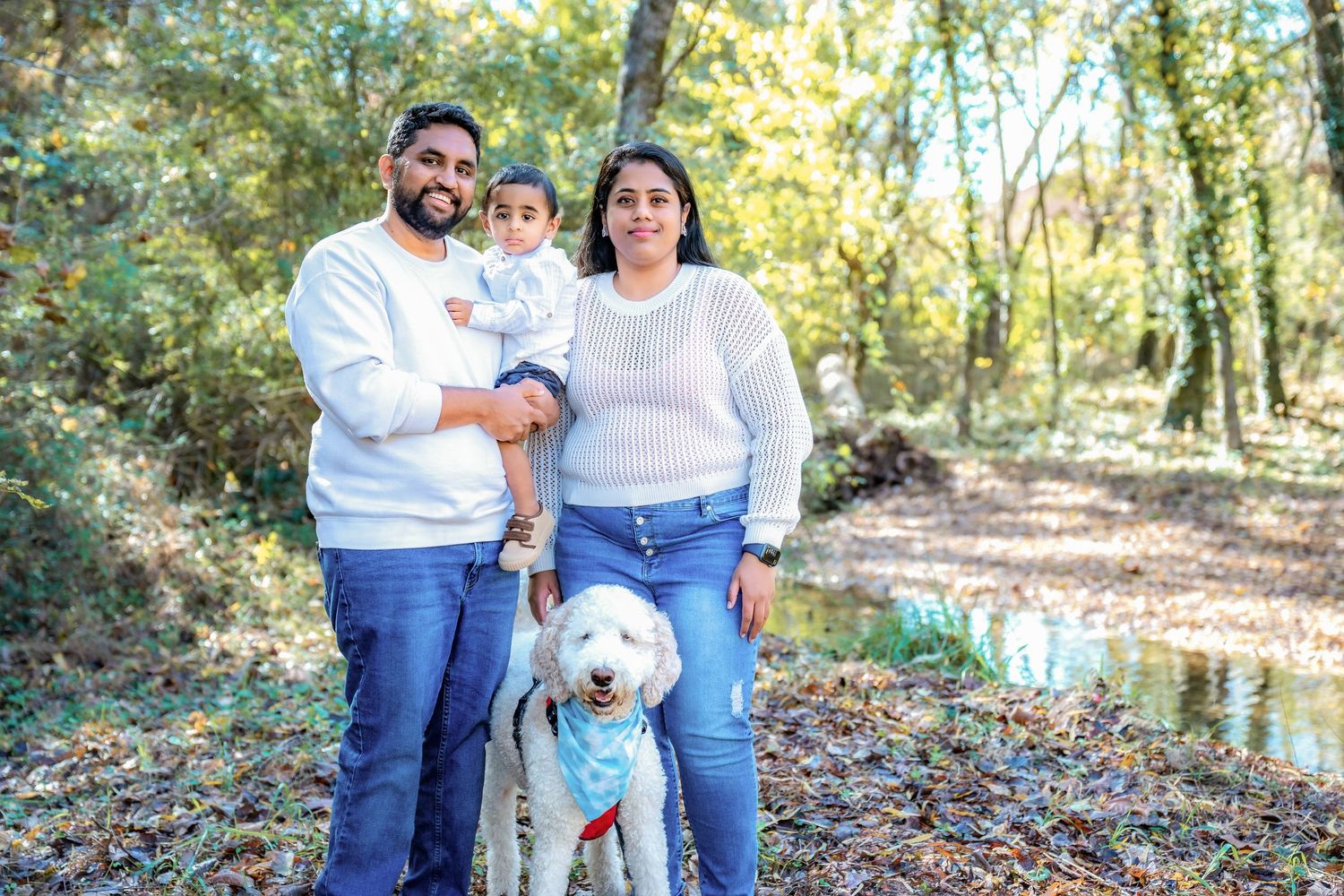 Family posing together outdoors in a wooded autumn setting with fallen leaves.