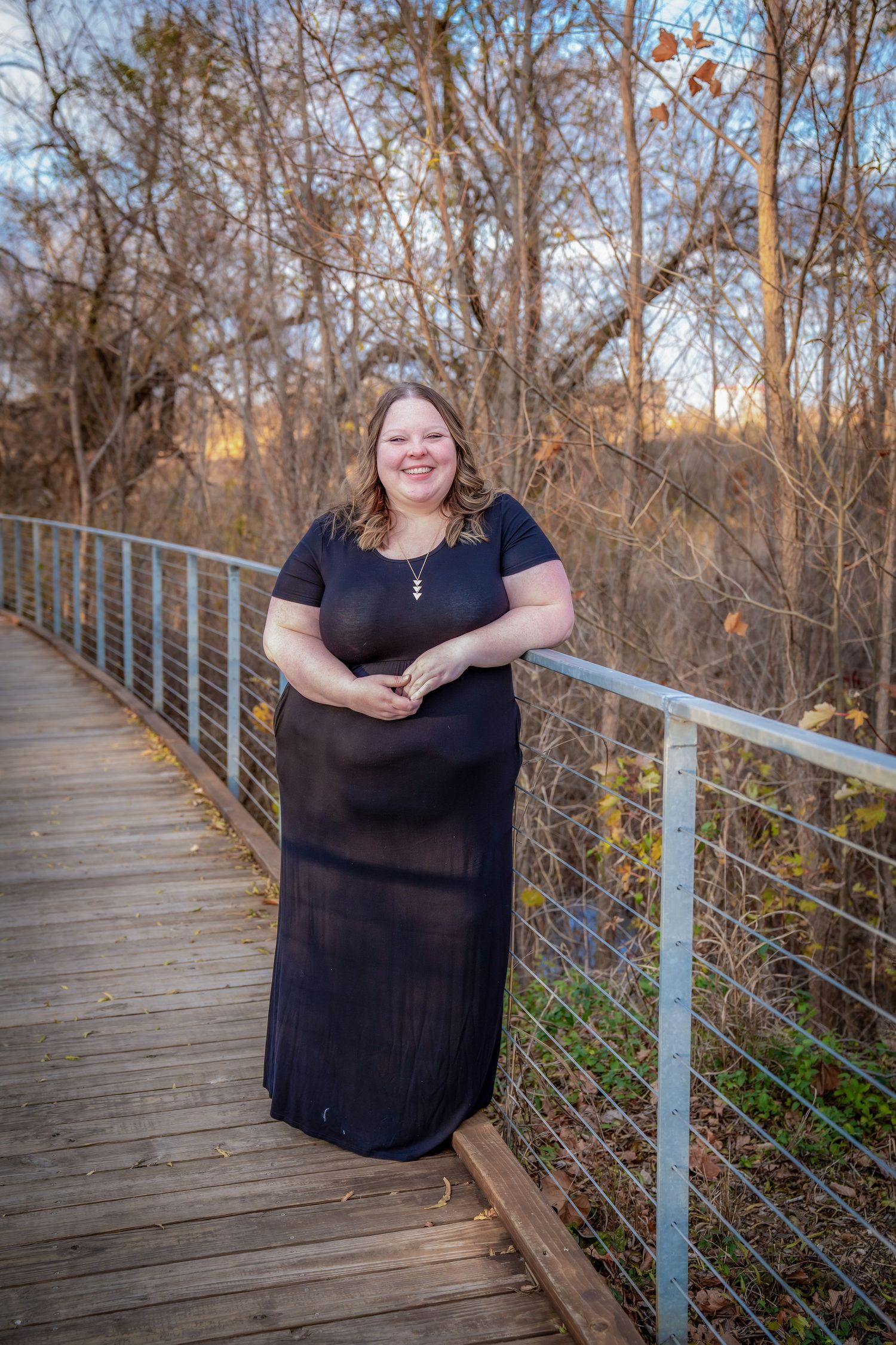 A person in a black dress stands on a wooden boardwalk with metal railings during autumn.