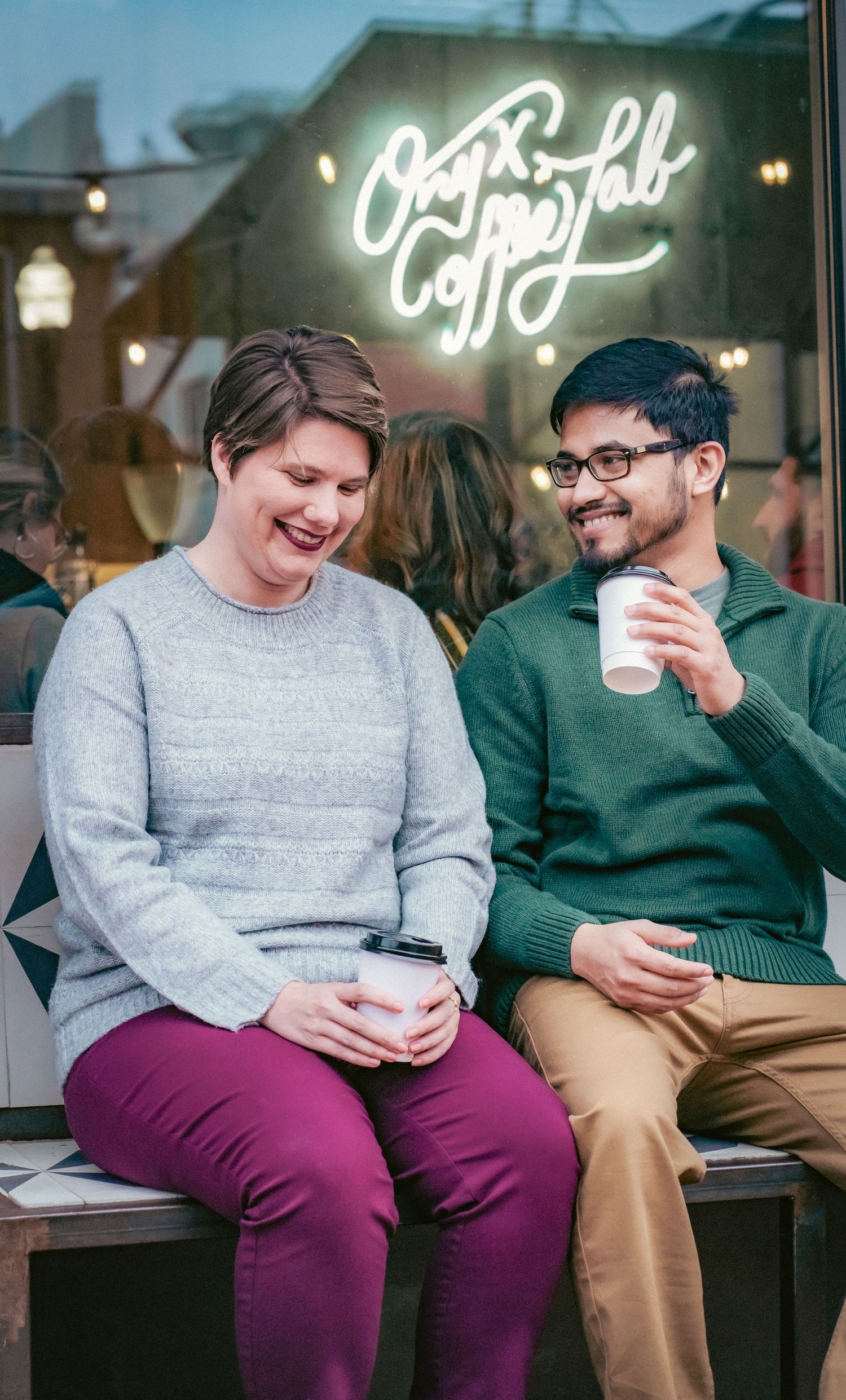 Two people enjoying coffee and conversation on a bench outside a cafe with neon signage.