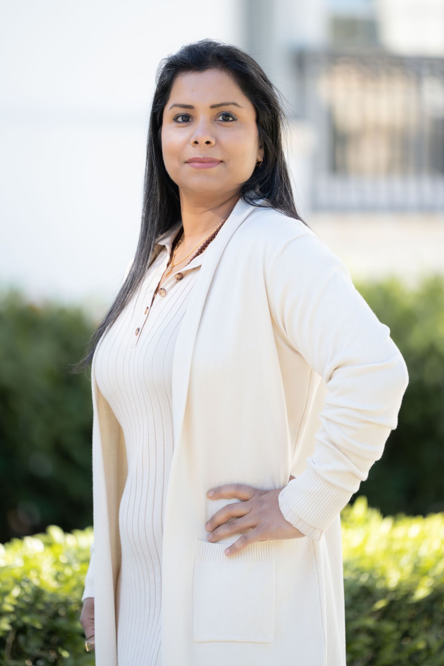 Professional portrait of someone in a cream-colored cardigan standing outdoors against a blurred garden background.
