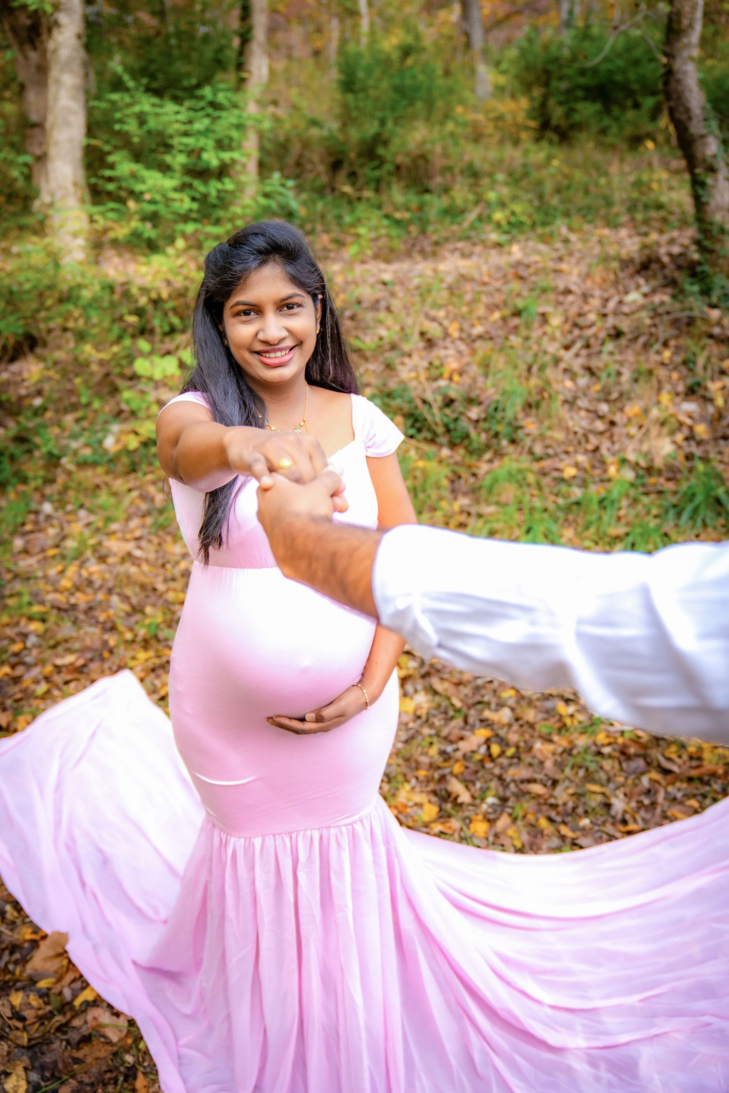 A flowing pink maternity gown billows gracefully in a wooded autumn setting.