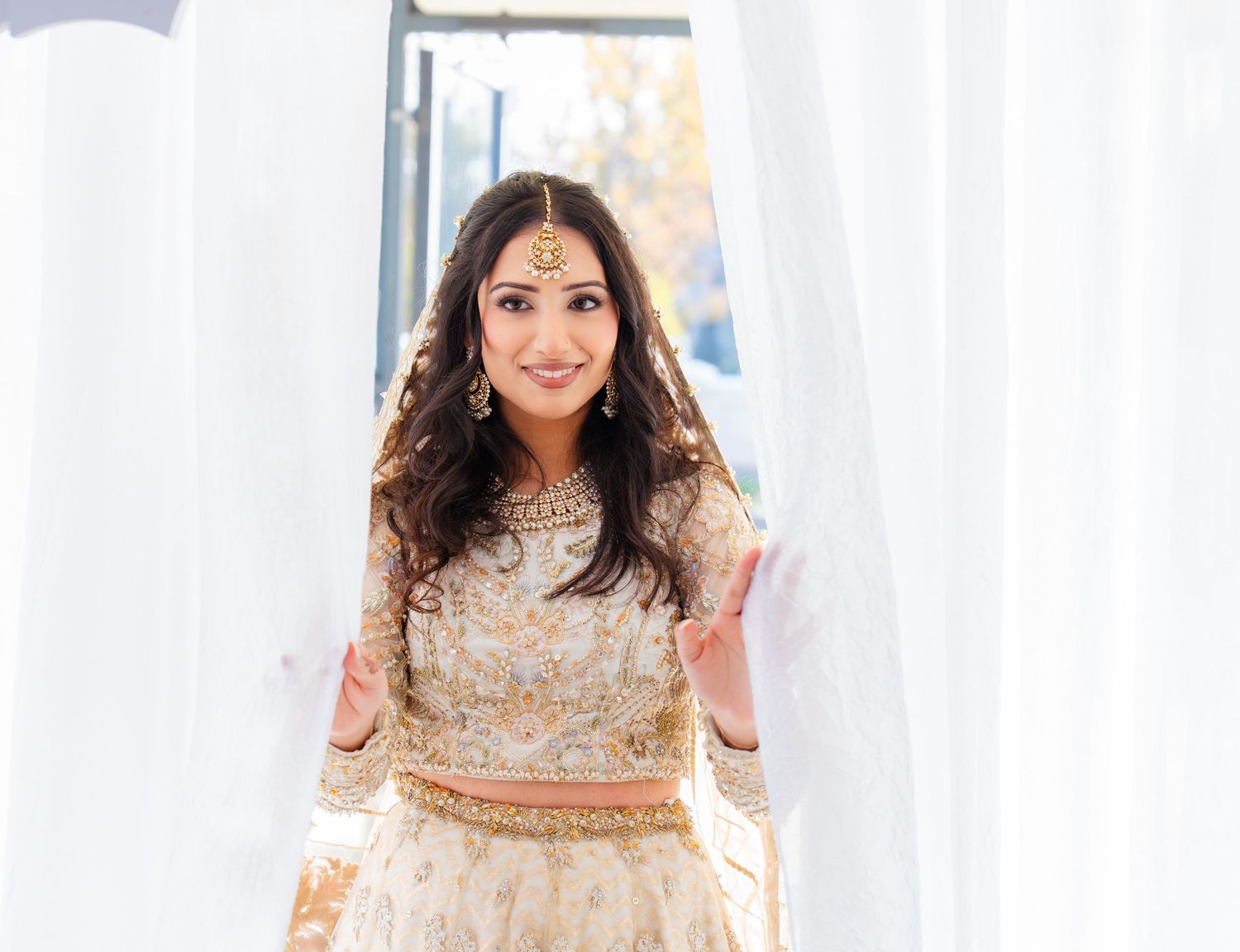 Bride in embellished cream and gold South Asian wedding attire poses against white drapes.