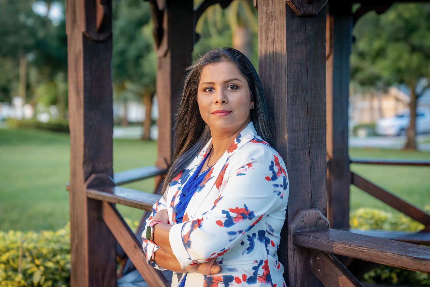 A person in a white floral dress poses in front of a wooden gazebo on a sunny day.