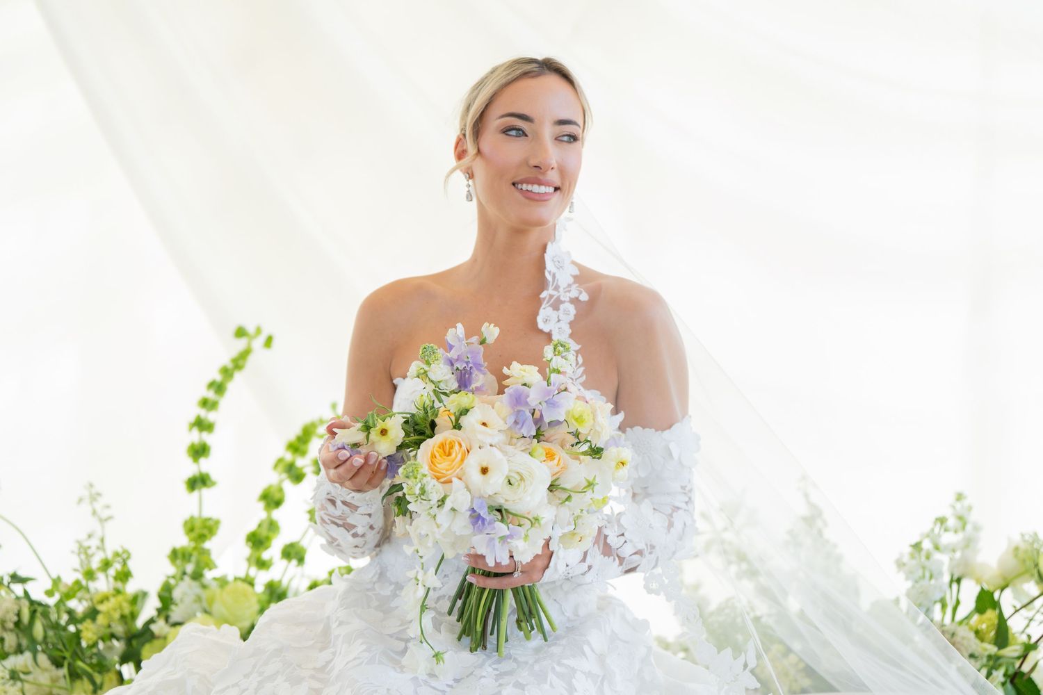 Bride in a white lace gown holds a pastel bouquet against a backdrop of greenery.