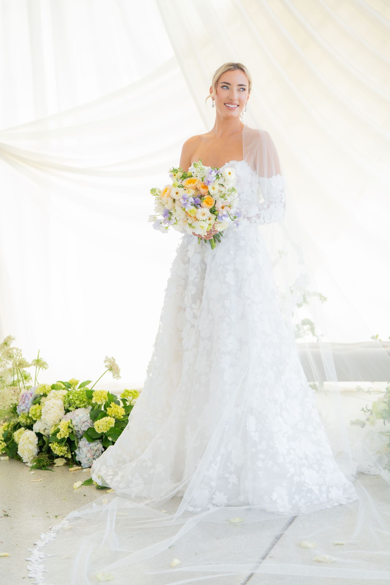 Bride in white lace gown holds pastel bouquet against ethereal white backdrop.