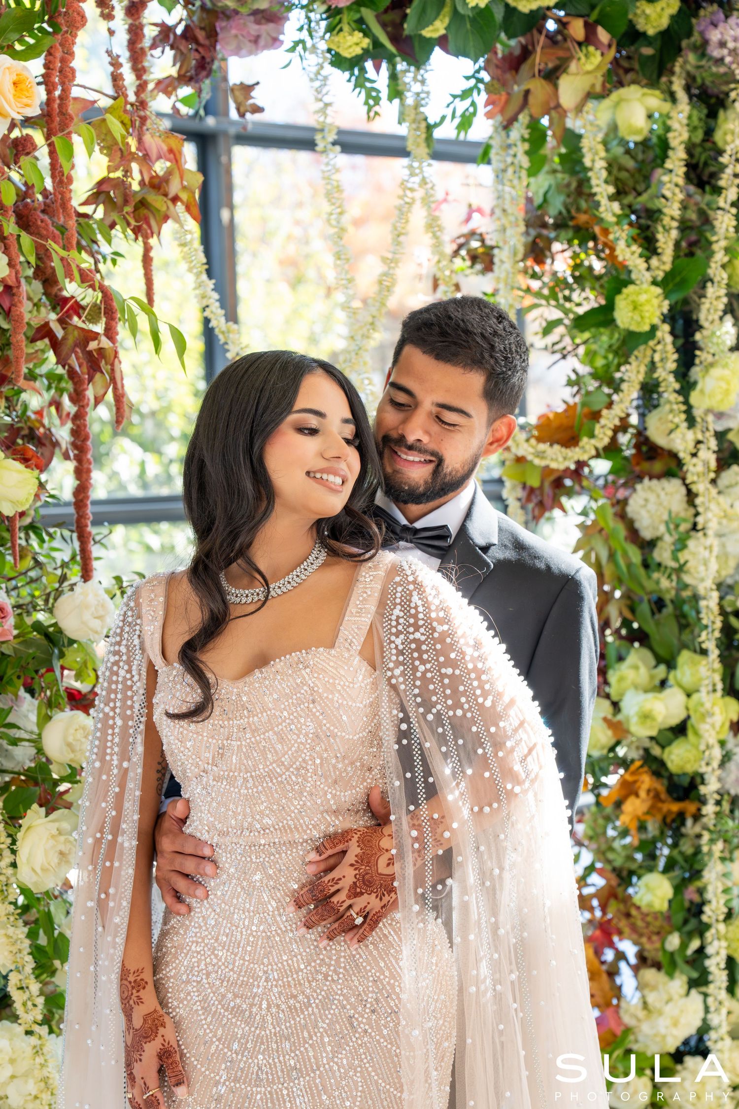 Couple embraces under a lush garden-inspired floral installation in formal wedding attire.