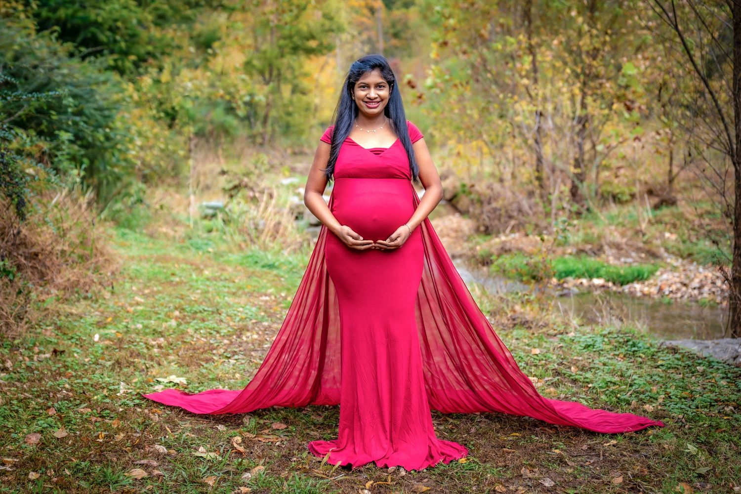 A flowing red maternity gown creates a dramatic silhouette against an autumn forest backdrop.
