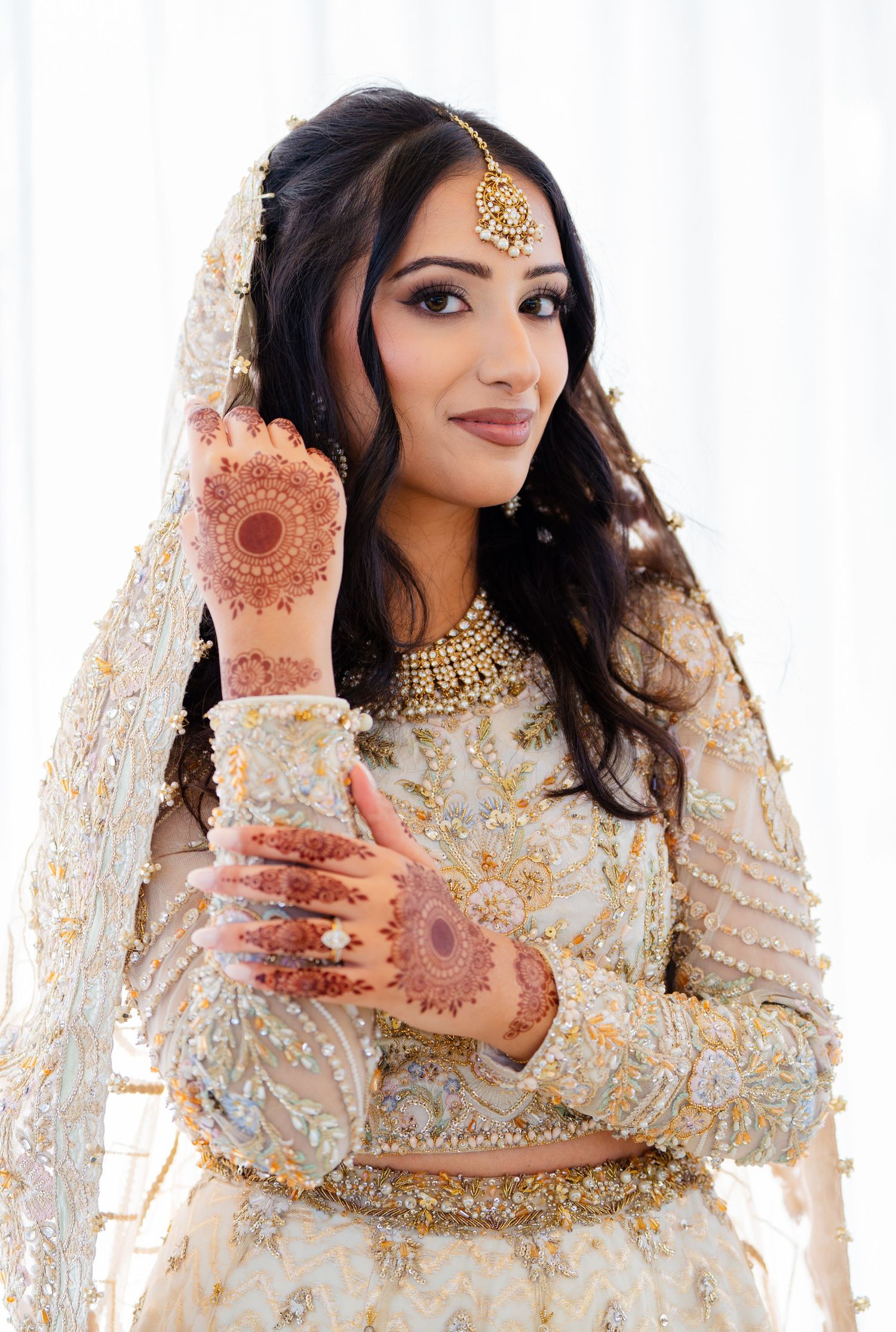 Traditional bride in cream and gold attire displaying intricate henna patterns on hands.