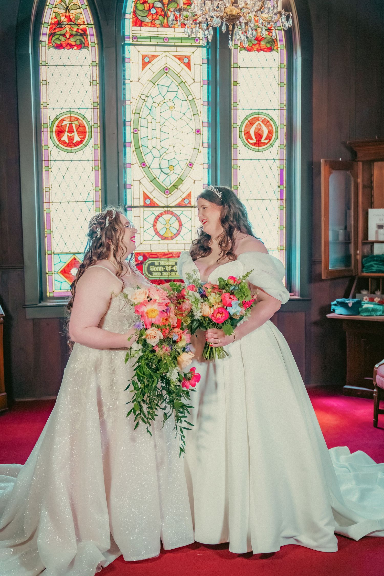 Two people in white wedding dresses pose by stained glass windows with colorful bouquets.
