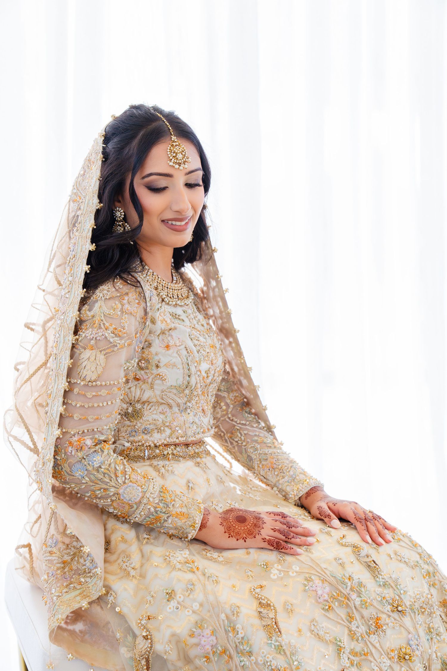 Bride in embellished cream lehenga smiling while seated against white backdrop.