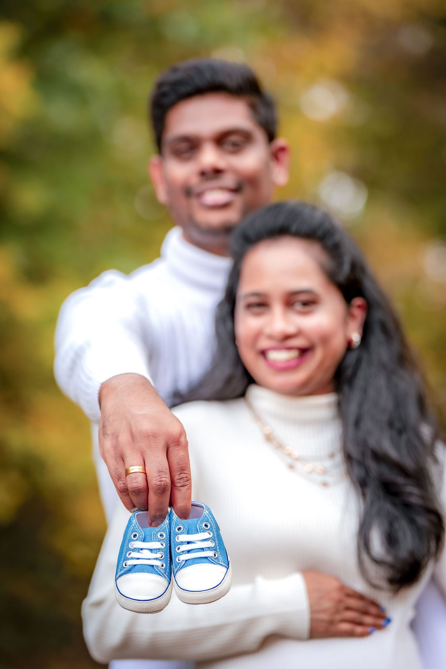 A couple holds tiny blue baby shoes while embracing outdoors in an autumn setting.