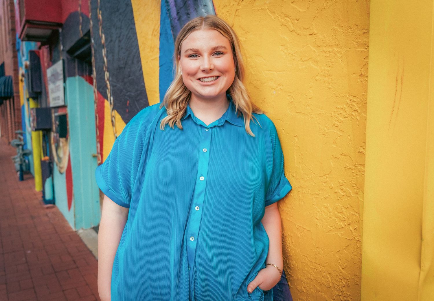 A bright turquoise outfit styled with blue sneakers against a vibrant yellow and red wall background.