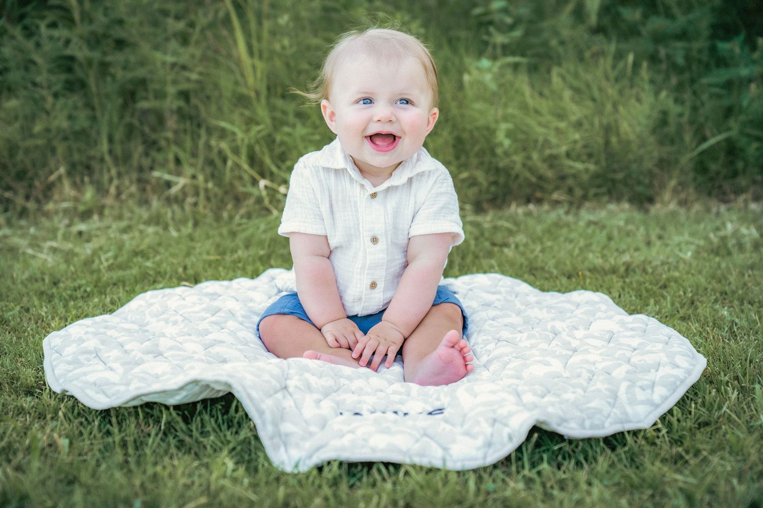 A baby sits on a white blanket in a grassy outdoor setting during a summer photoshoot.