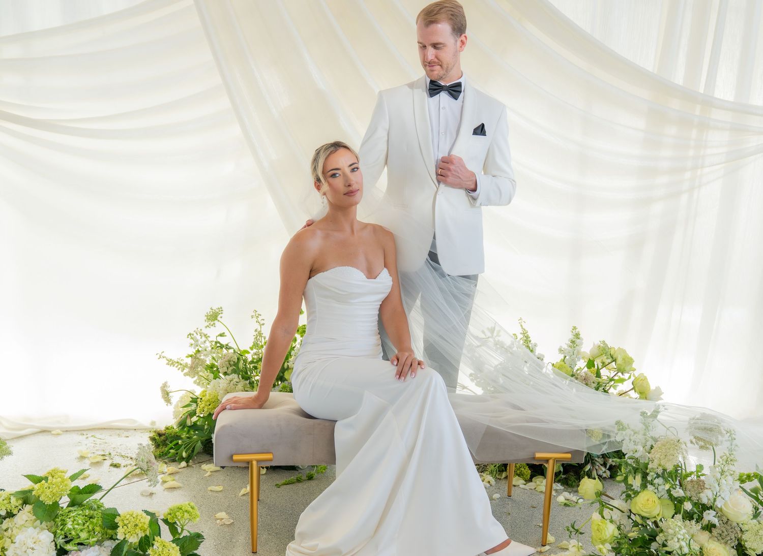 Elegant wedding portrait in a white draped setting with gold accents and greenery.
