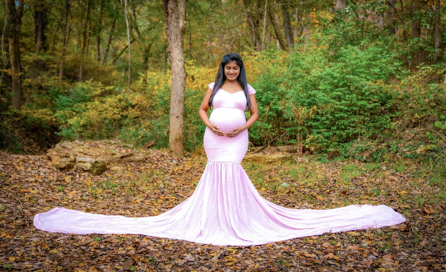A pink maternity dress trails elegantly across fallen autumn leaves in a forest setting.