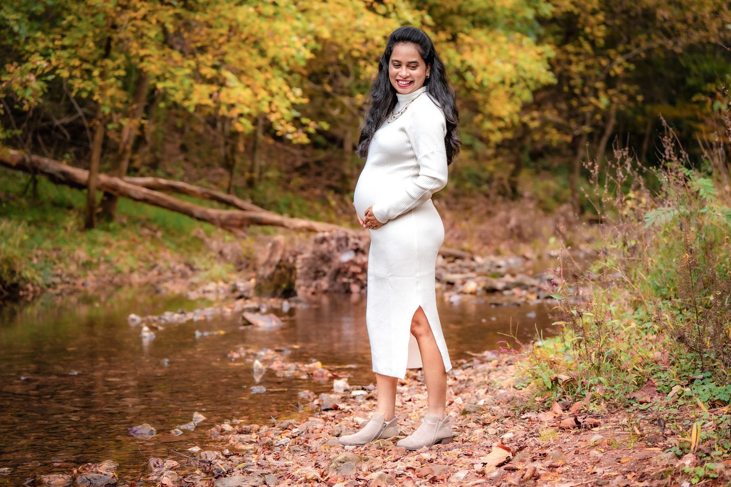 A woman in white poses gracefully along a wooded path with autumn leaves scattered on the ground.