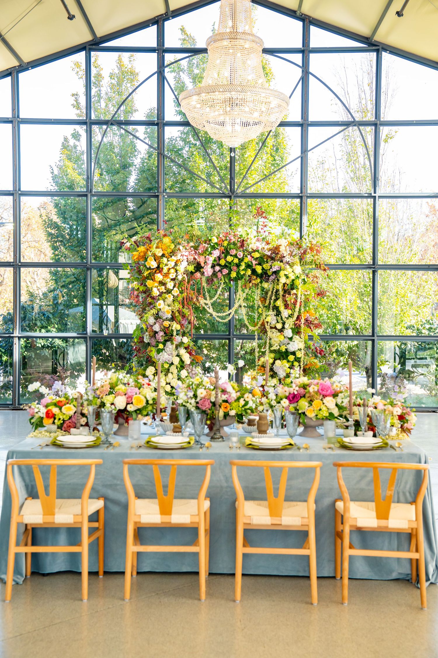 Elegant reception setup in glass conservatory with wooden chairs and hanging floral arrangements.