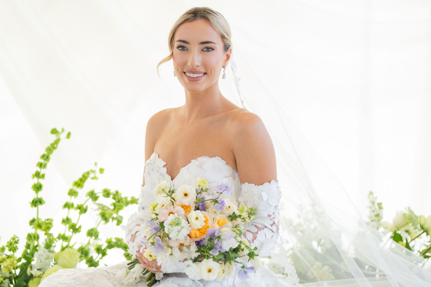 Bride in strapless white gown holds spring bouquet against white backdrop with green accents.