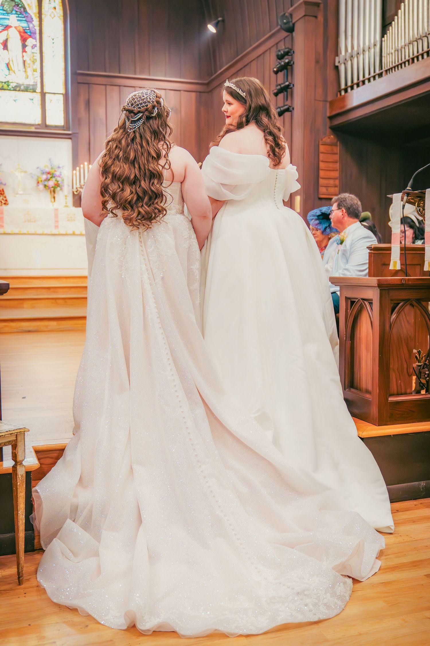 Two brides in elegant white dresses dance together on wooden church floors.