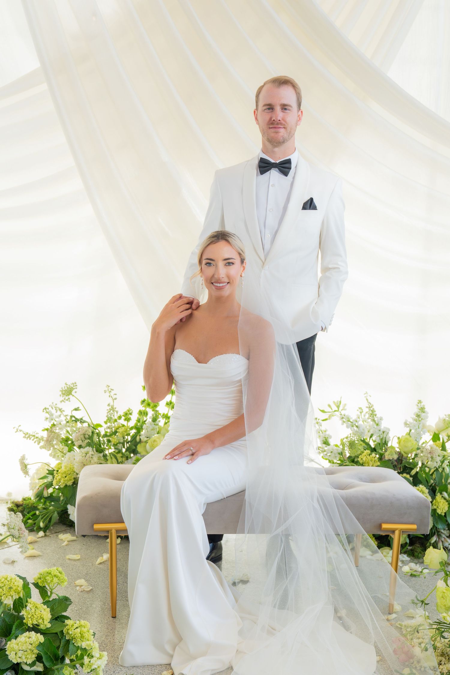 Elegant wedding portrait in a white draped setting with lush green floral arrangements.