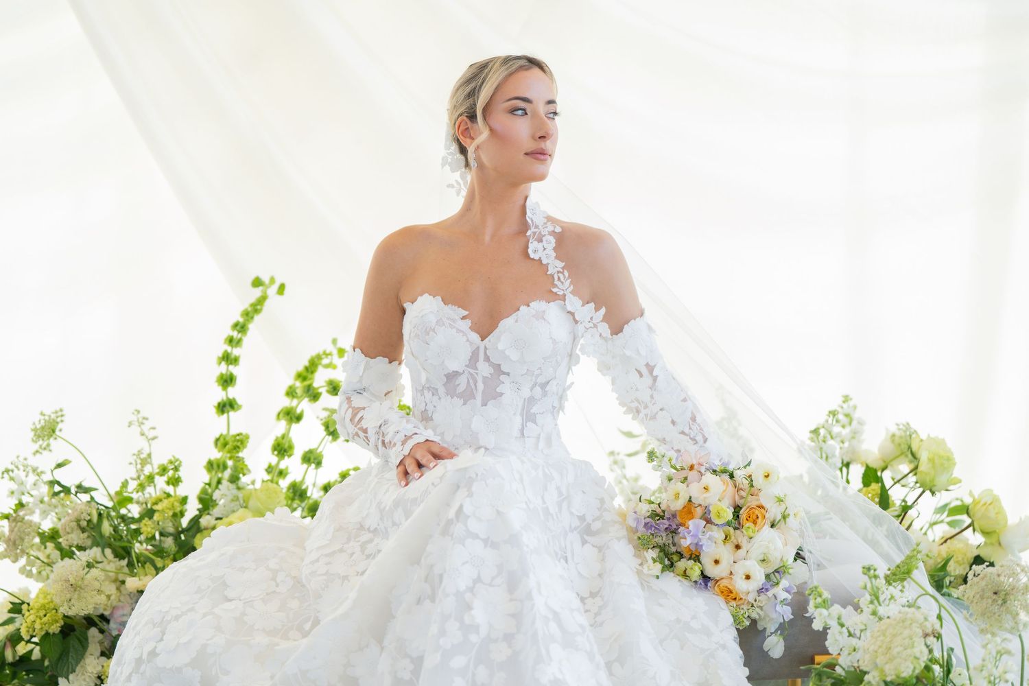 Bride in detailed white gown with off-shoulder design holds pastel floral bouquet.